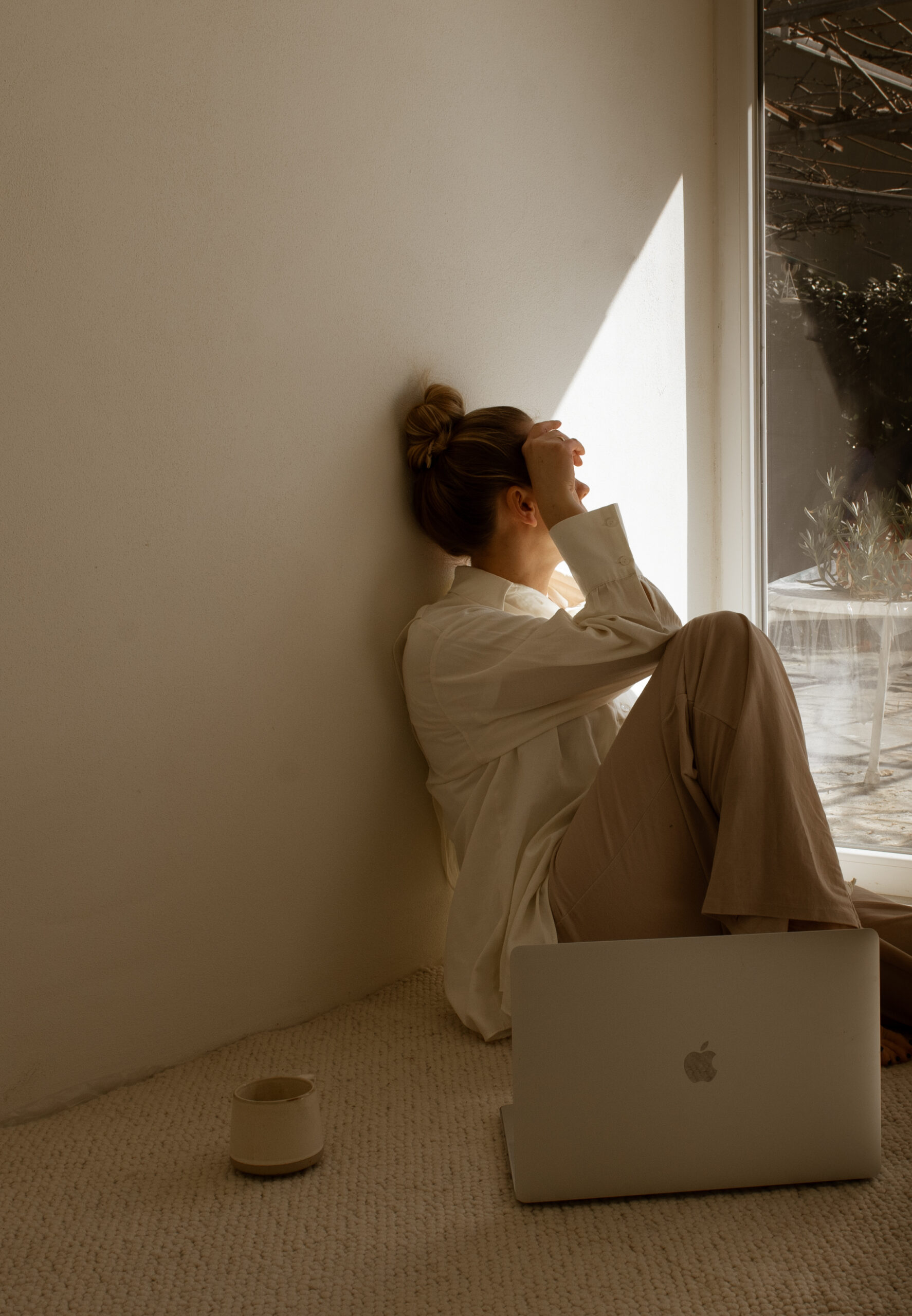 Woman sitting on the floor in front of a window with her hand on her head, looking overwhelmed